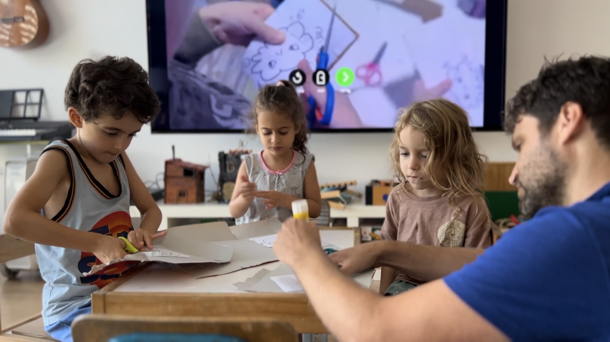 A parent and three children cutting and gluing a Catbears craft together at the table, with the activity playing on the screen behind them