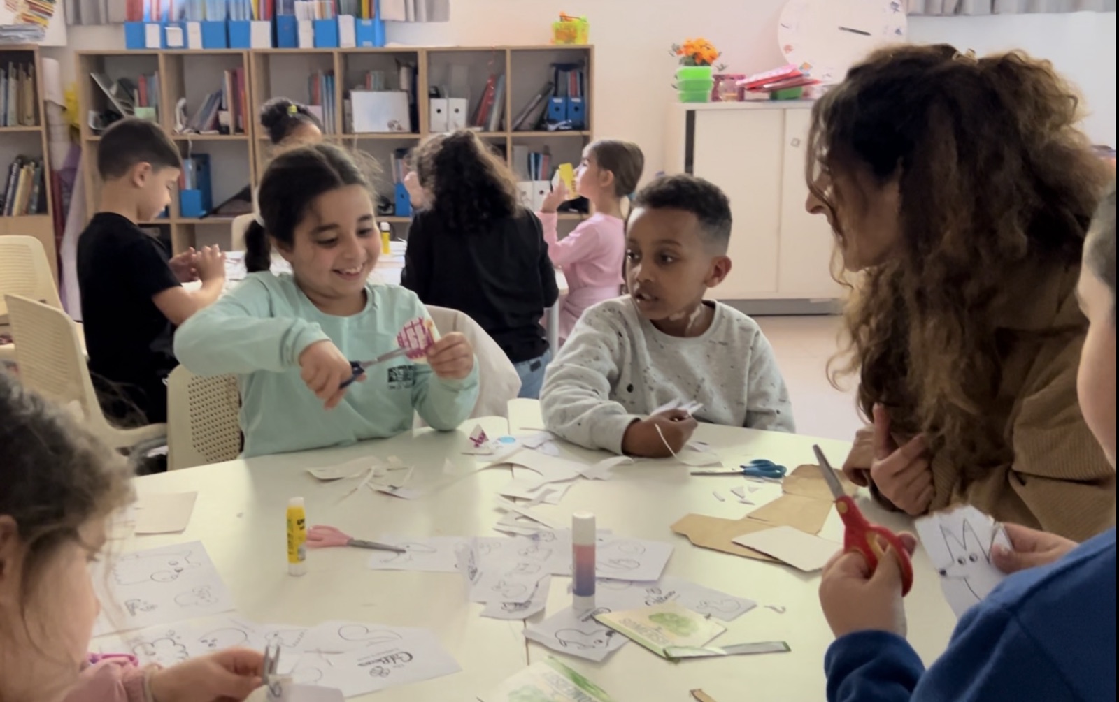A teacher leaning in at a classroom table while children cut out Catbears puppets together